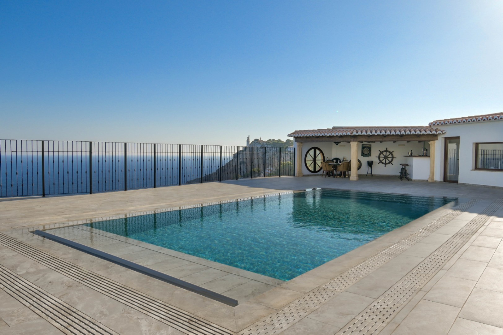 Piscina de borde infinito en terraza de gres con vistas al mar y al faro. Dispone de un porche cubierto con vigas de madera, zona de comedor y detalles náuticos.