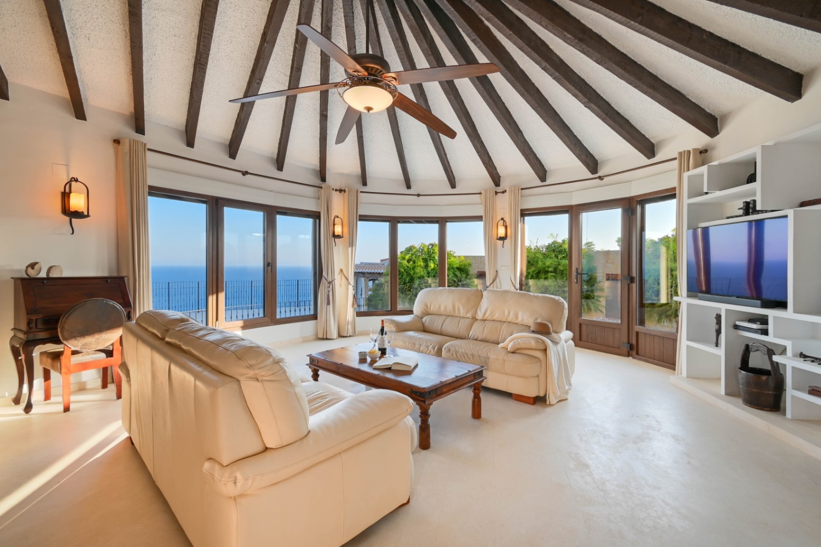 Salón circular con techo de vigas de madera radiales, sofás de cuero crema y amplios ventanales con vistas panorámicas al mar.