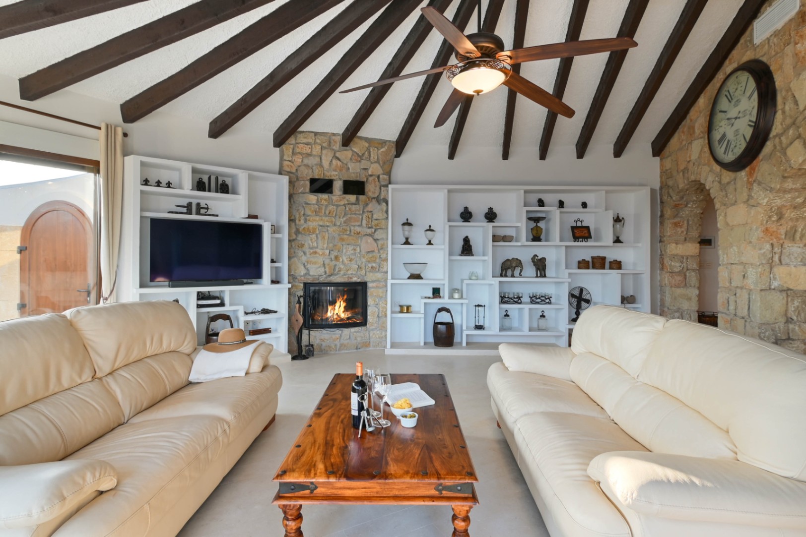 Salón con techo de vigas de madera, chimenea de piedra encendida y estanterías blancas integradas. Incluye sofás de cuero crema y una mesa de centro de madera rústica.