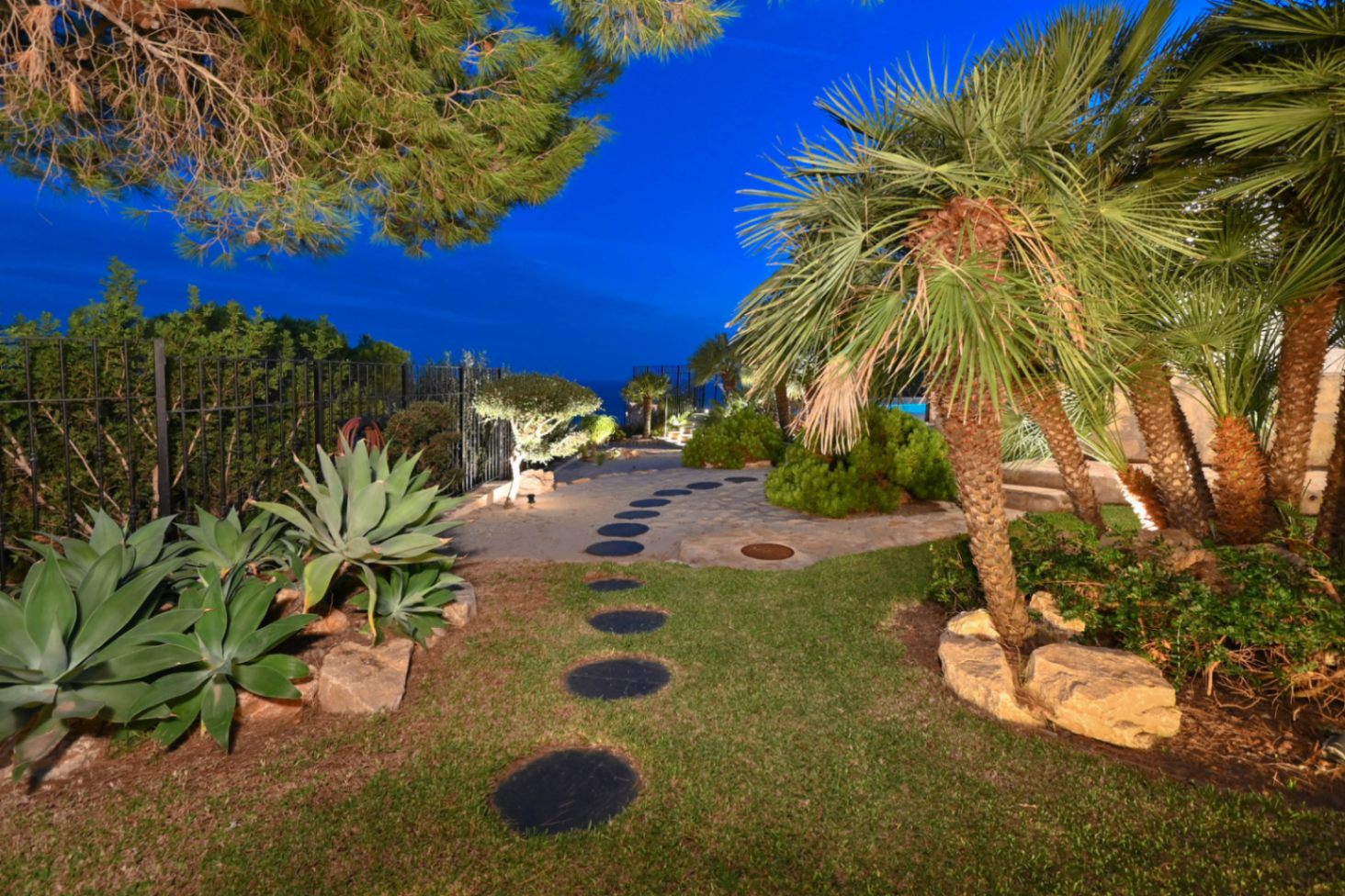 Sendero de piedras circulares en un jardín mediterráneo con palmeras, pitas e iluminación exterior integrada bajo el cielo del atardecer.