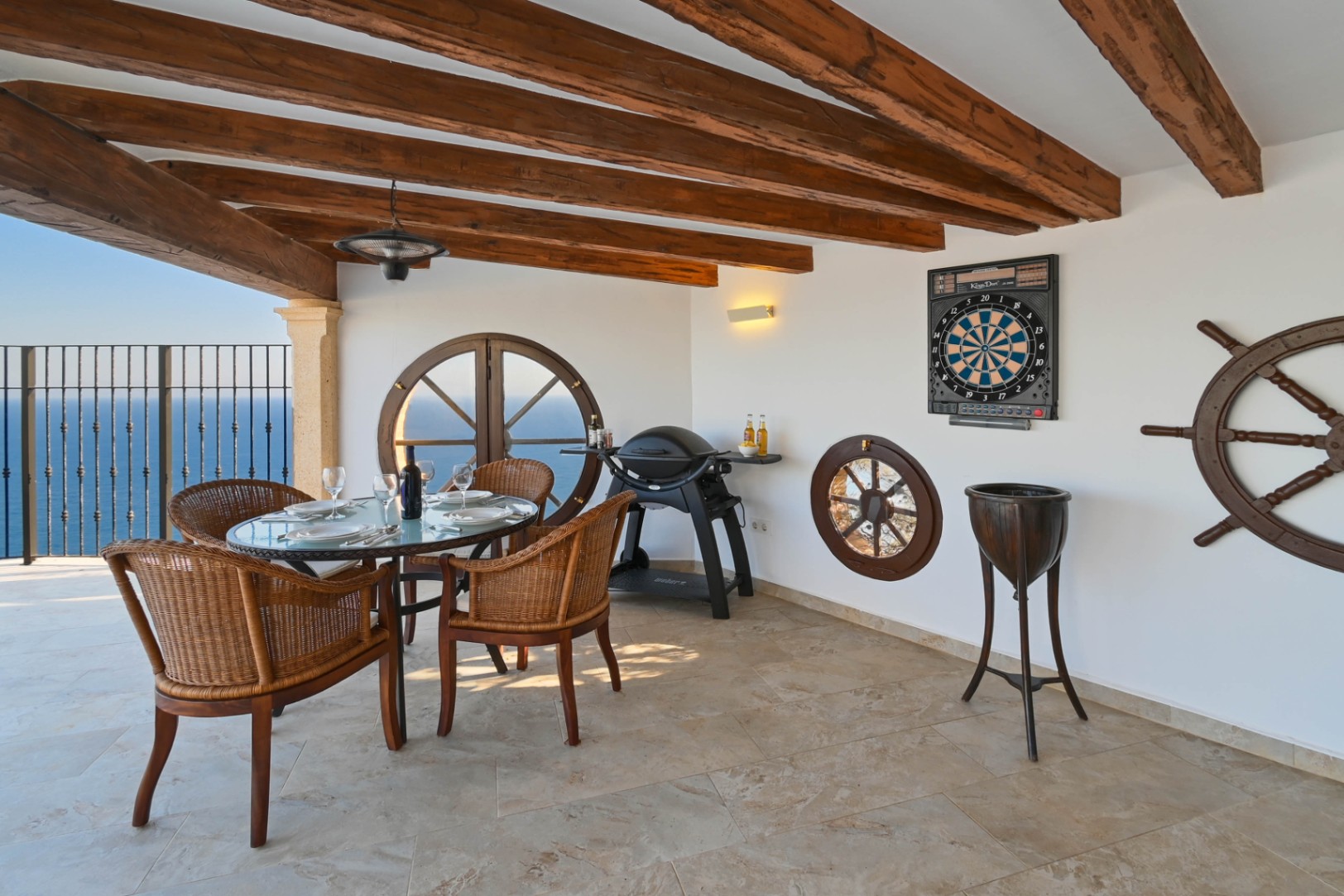 Terraza cubierta con vigas de madera, ventanas circulares tipo buey y vistas al mar. Dispone de comedor de mimbre, barbacoa de gas y elementos decorativos náuticos.
