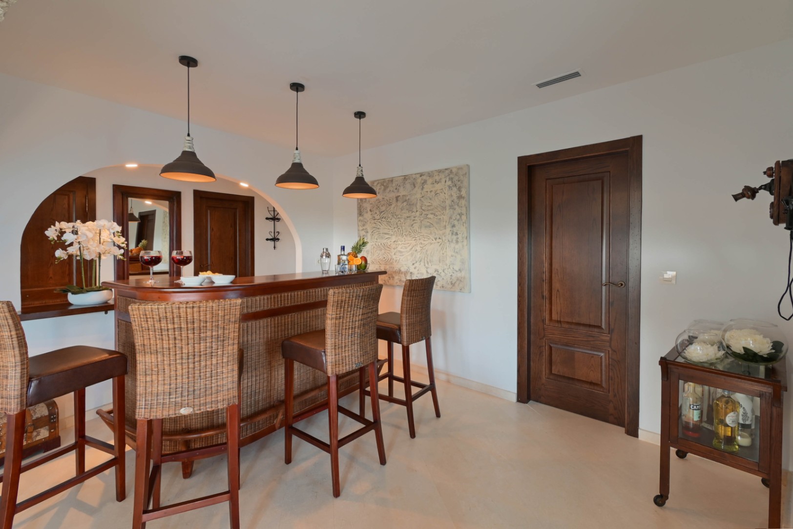 Zona de bar interior con barra de madera curva y detalles de mimbre, taburetes altos, lámparas colgantes industriales y puertas de madera oscura de estilo clásico.