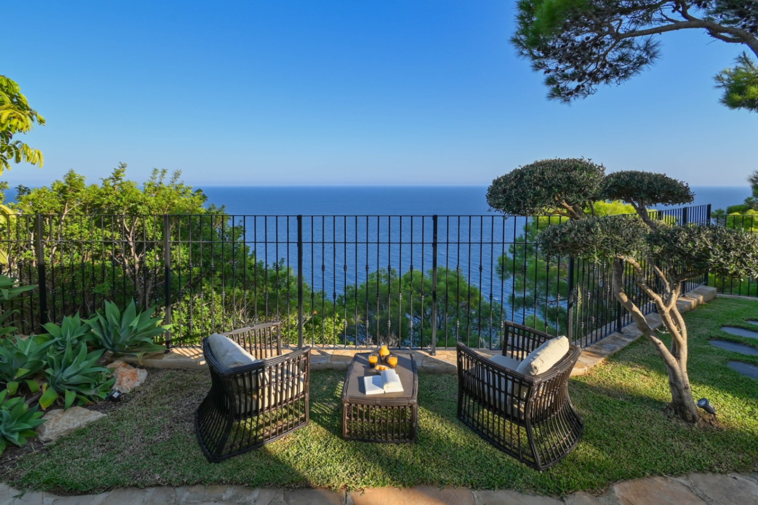 Zona de estar con sillones de mimbre oscuro y mesa sobre césped, con vistas panorámicas al mar tras una barandilla de hierro forjado.