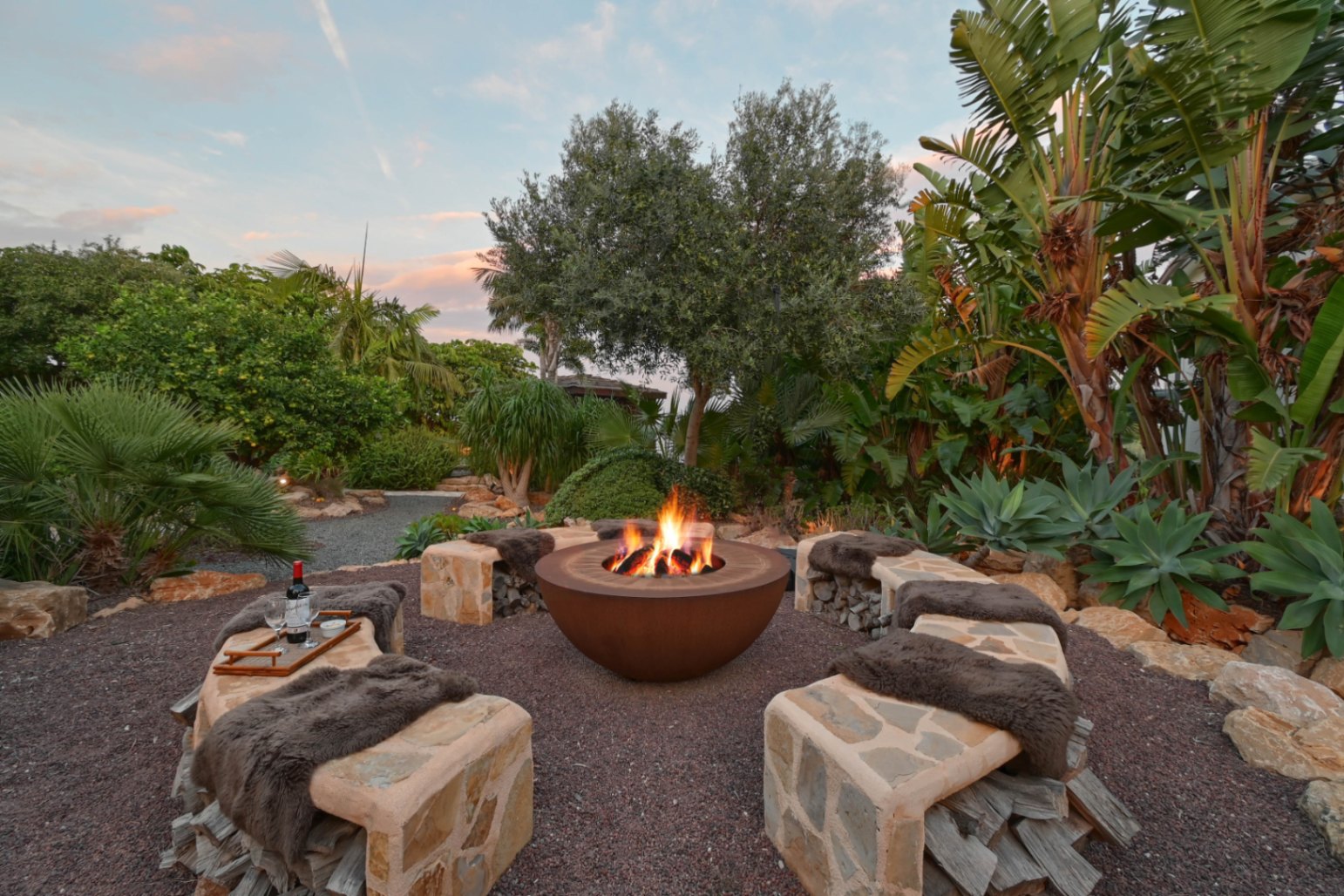 Zona de fuego exterior con brasero de acero corten, bancos de piedra con leñero integrado y vegetación tropical exuberante bajo el cielo del atardecer.