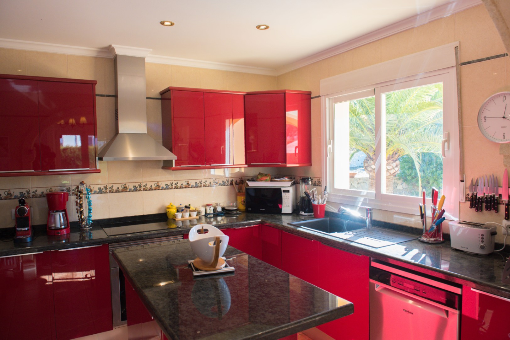 Cocina funcional con armarios rojos de alto brillo, encimeras de granito negro y electrodomésticos de acero inoxidable. Incluye una ventana con vistas a la vegetación exterior.