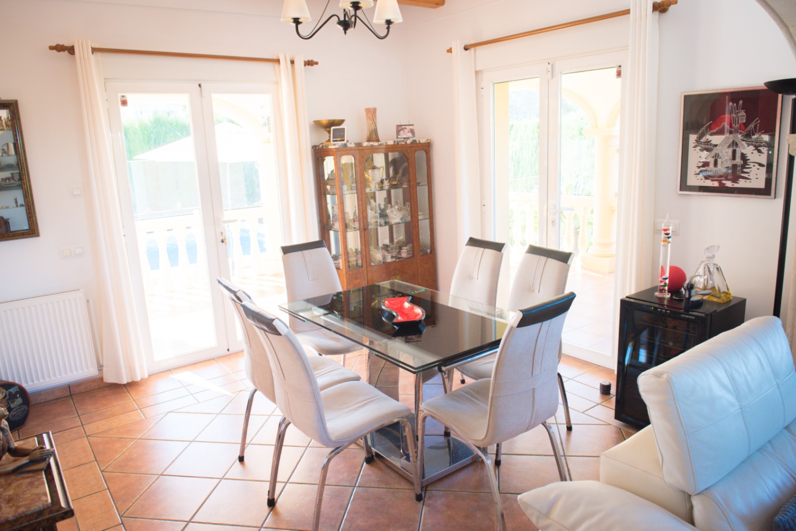 Comedor con mesa de cristal y sillas blancas sobre suelo de terracota. Puertas dobles acristaladas iluminan el espacio y conectan con la terraza.