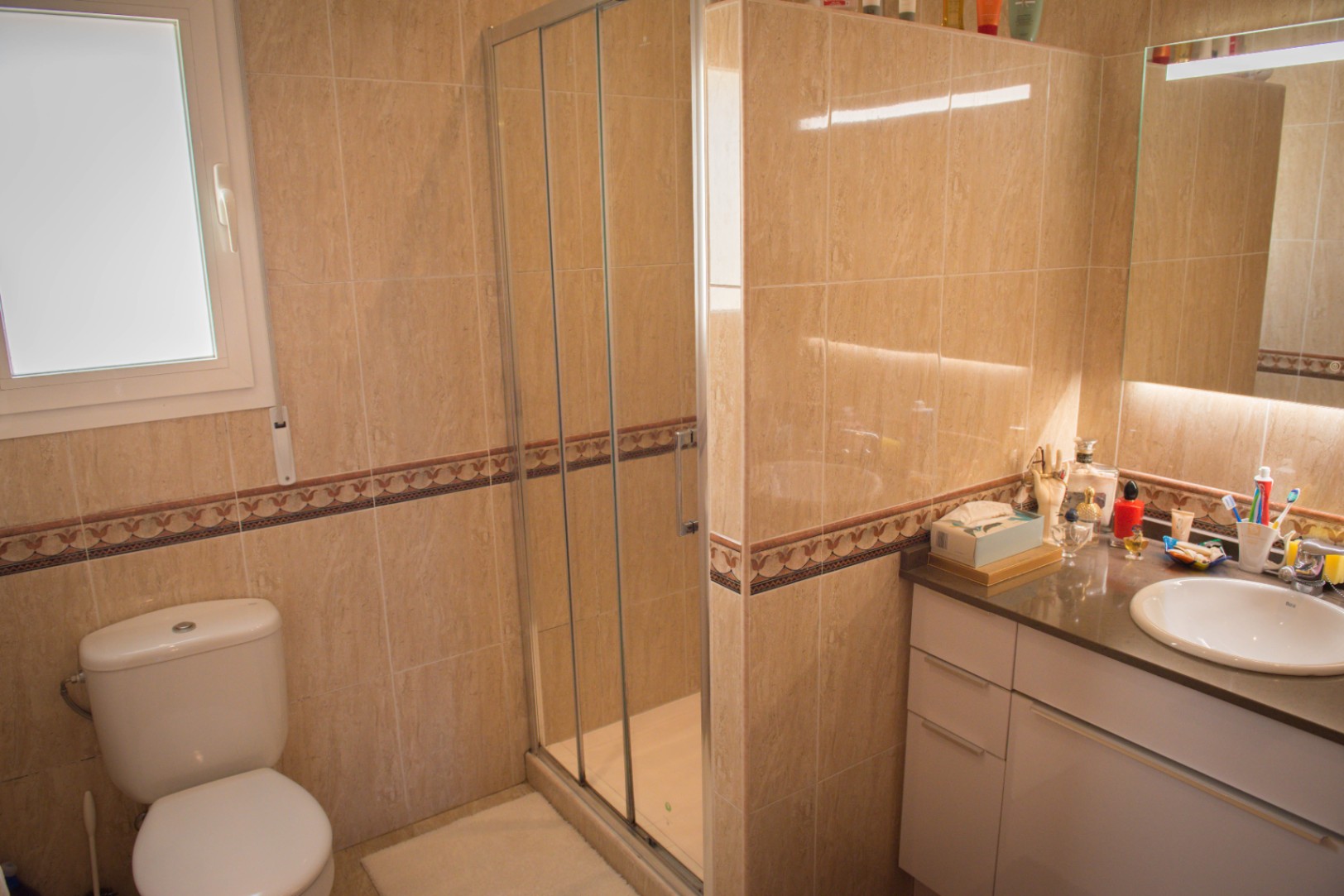 Cuarto de baño con azulejos beige y cenefa decorativa, equipado con ducha de cristal, inodoro blanco y lavabo con espejo retroiluminado.