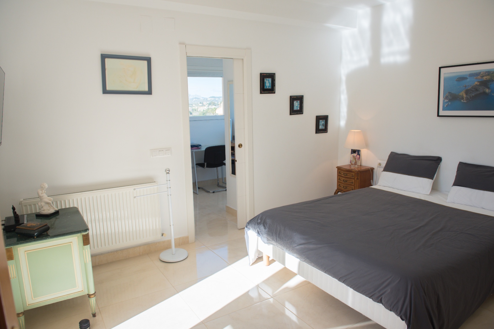 Dormitorio con suelos de baldosa clara, cama doble y mobiliario clásico. Una puerta abierta conecta con un despacho privado luminoso con vistas.