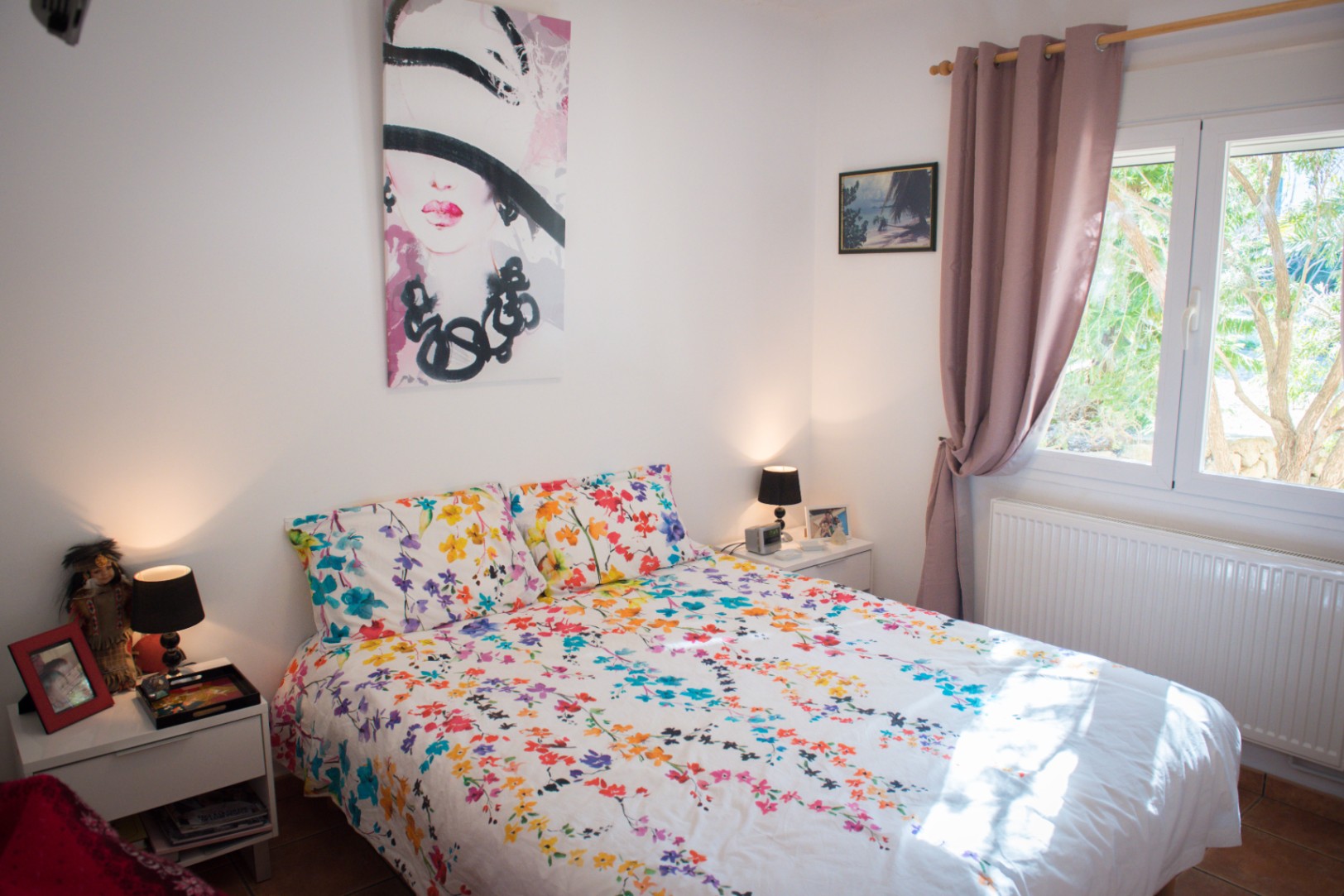 Dormitorio luminoso con ropa de cama floral, un gran cuadro de moda, mesitas de noche blancas con lámparas negras y ventana con cortinas malva hacia el exterior.