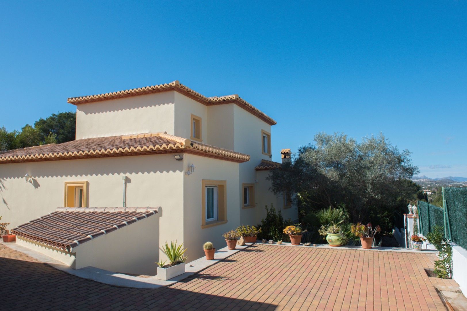 Exterior de villa mediterránea con fachadas color crema, tejados de teja cerámica y una amplia terraza de ladrillo decorada con plantas en macetas.