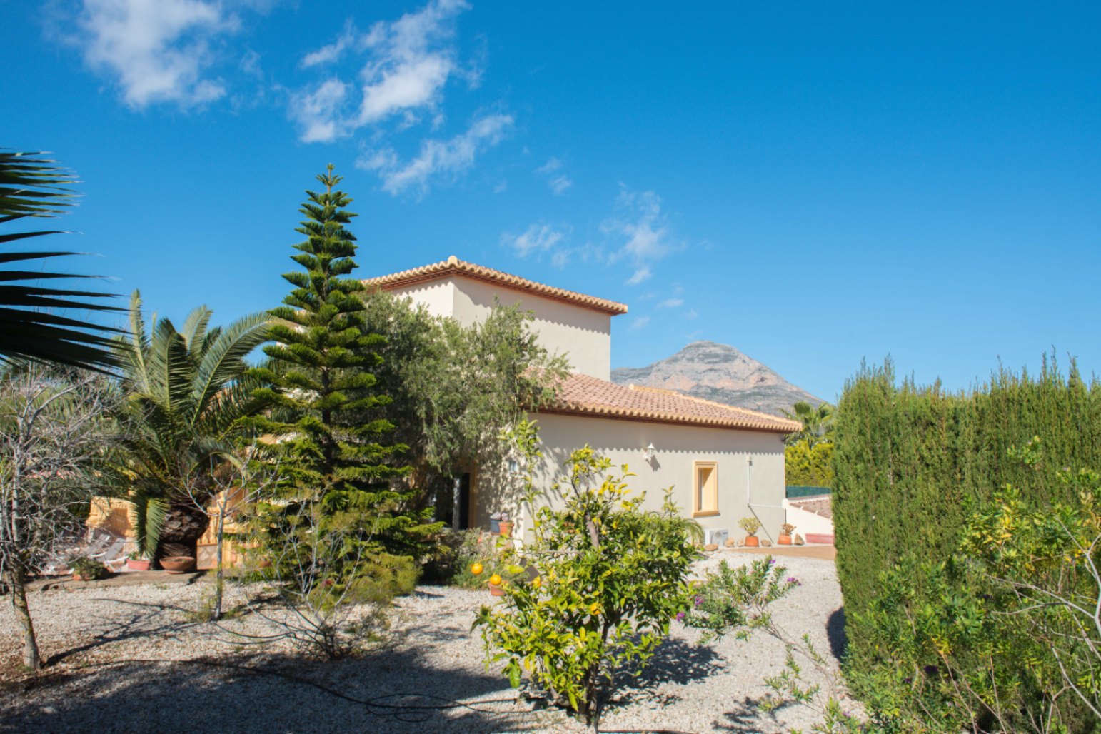 Exterior de villa mediterránea con tejado de terracota, jardín de grava con cítricos, palmeras y setos de cipreses con vistas a la montaña.
