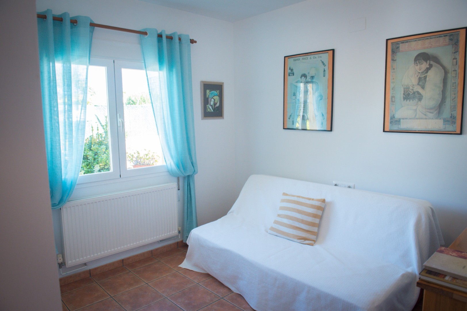Habitación luminosa con suelo de baldosas de terracota, sofá blanco con cojín a rayas y ventana con cortinas turquesas que aporta luz natural.