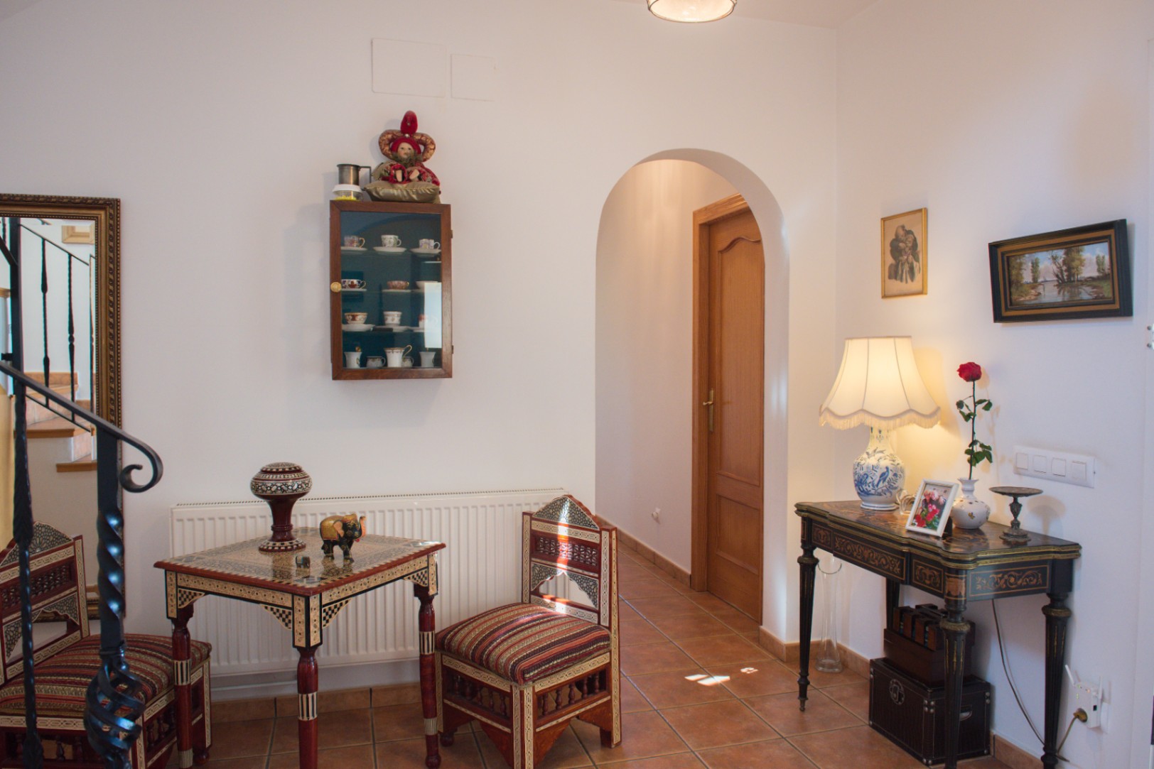 Recibidor con suelos de terracota, un arco de paso y mobiliario de madera taraceada junto a una consola decorativa y vitrina de pared.