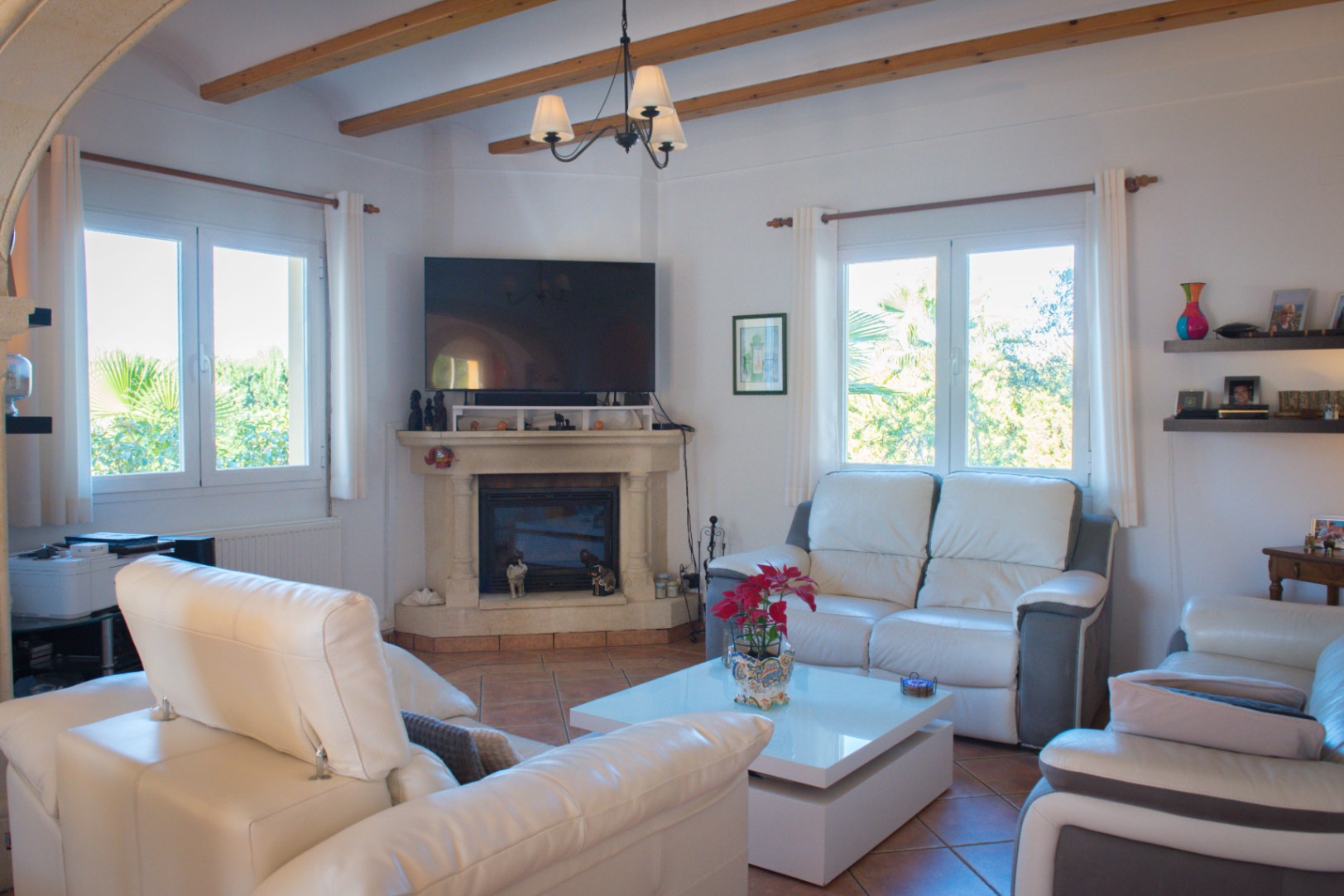 Salón con vigas de madera vistas, chimenea de piedra en esquina con televisión integrada, sofás de cuero blanco y suelo de baldosas de terracota.
