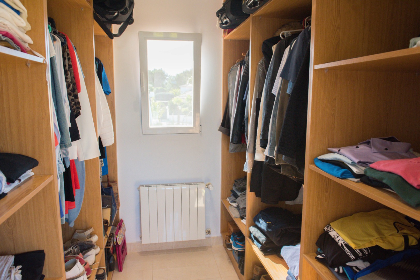 Vestidor con estanterías de madera de roble, barras para colgar y baldas abiertas. Incluye una ventana central para luz natural y un radiador de pared.