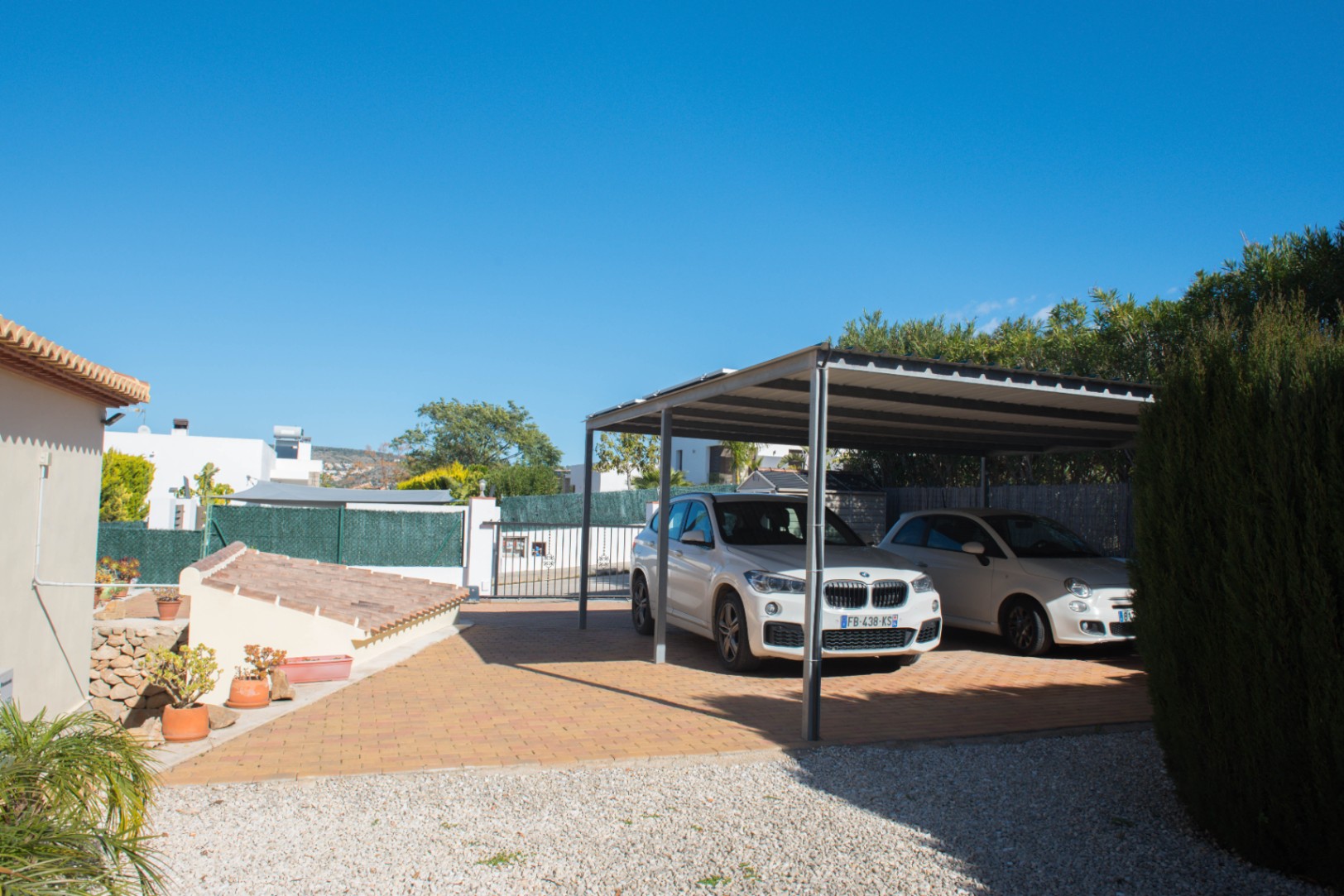 Zona de aparcamiento cubierta con estructura metálica, suelo de adoquines cerámicos y entorno de grava blanca con vegetación mediterránea.