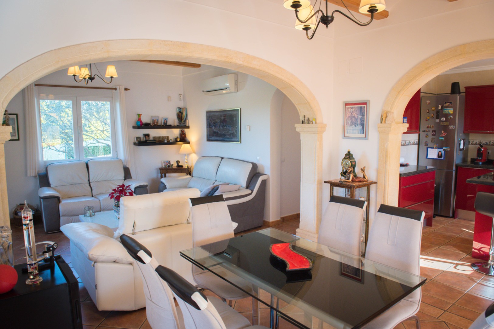 Zona de estar de planta abierta con arcos de piedra natural, comedor con mesa de cristal, sofás de cuero y cocina moderna con armarios rojos y suelo de terracota.