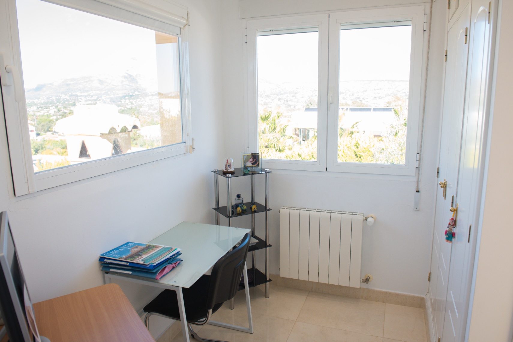 Zona de estudio luminosa con escritorio de cristal, armarios empotrados blancos y ventanas dobles con vistas despejadas a la montaña y el entorno urbano.