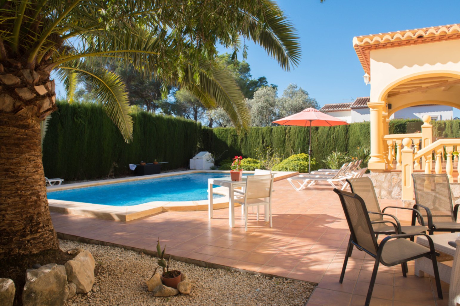 Zona de piscina privada con terraza de terracota, rodeada de setos altos y una palmera, junto a un porche con arcos de estilo mediterráneo.