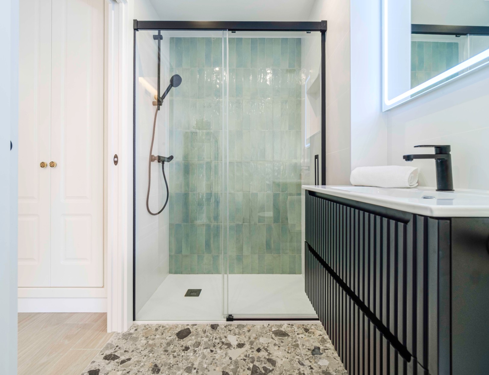 Baño moderno con ducha de azulejos verticales turquesa, grifería negra mate y mueble de lavabo alistonado con espejo retroiluminado.