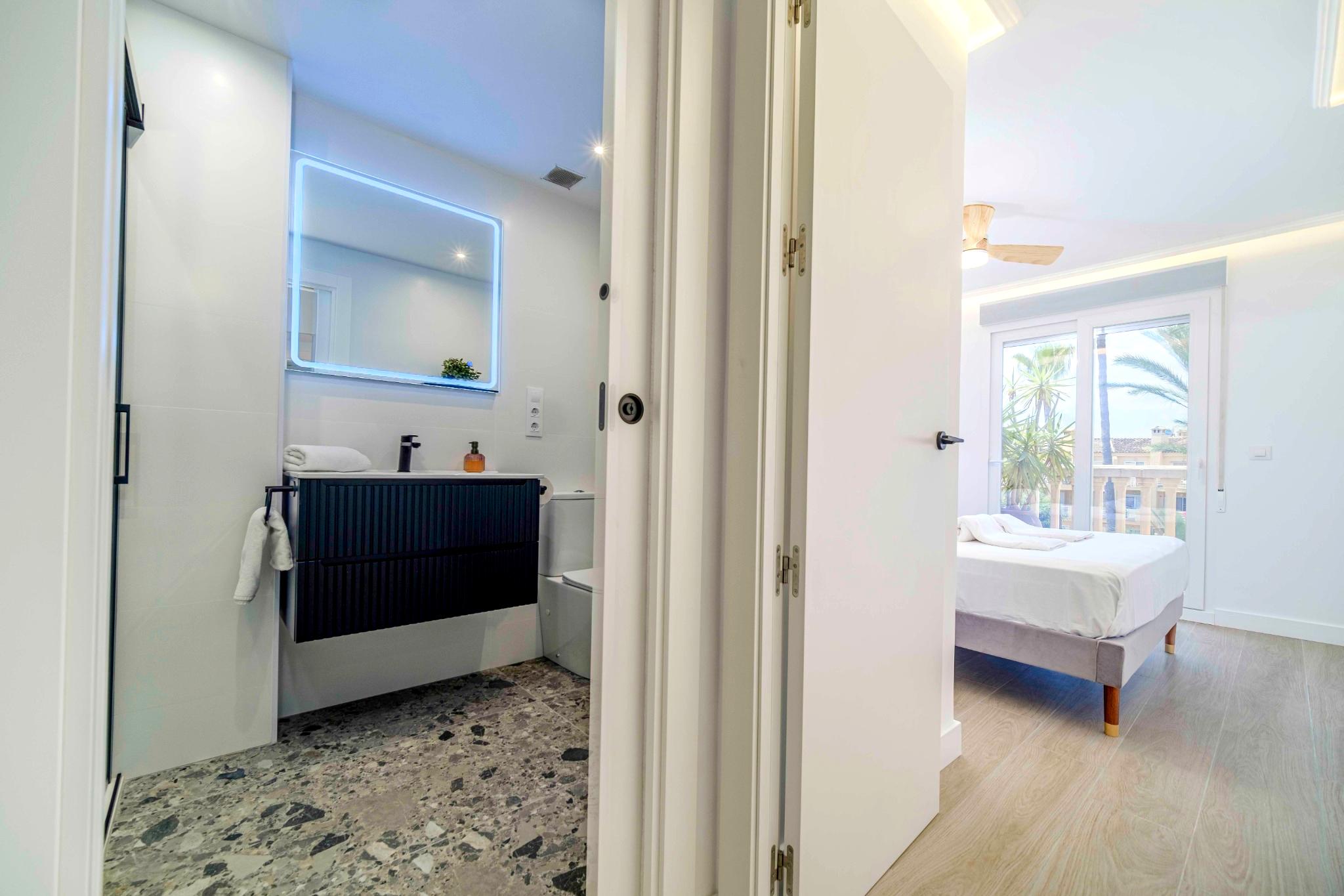Baño moderno con suelo de terrazo y mueble negro, conectado a un dormitorio luminoso con acceso a un balcón con vistas a las palmeras.