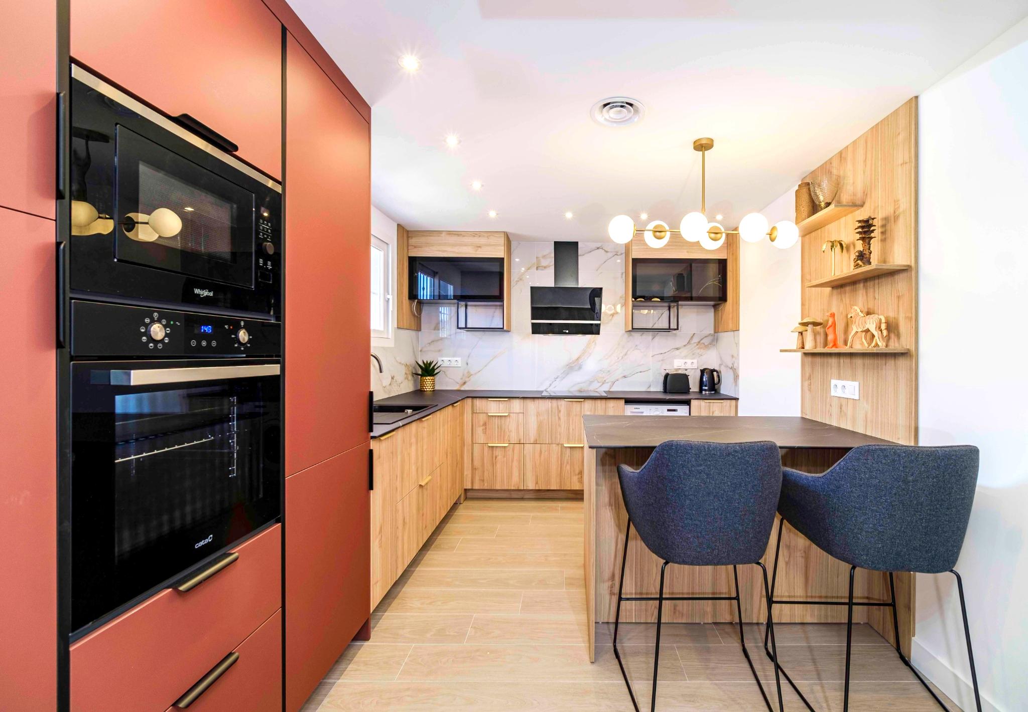 Cocina moderna con armarios en terracota y madera clara, encimera oscura, salpicadero de mármol y una isla con taburetes azules bajo una lámpara de globos dorada.