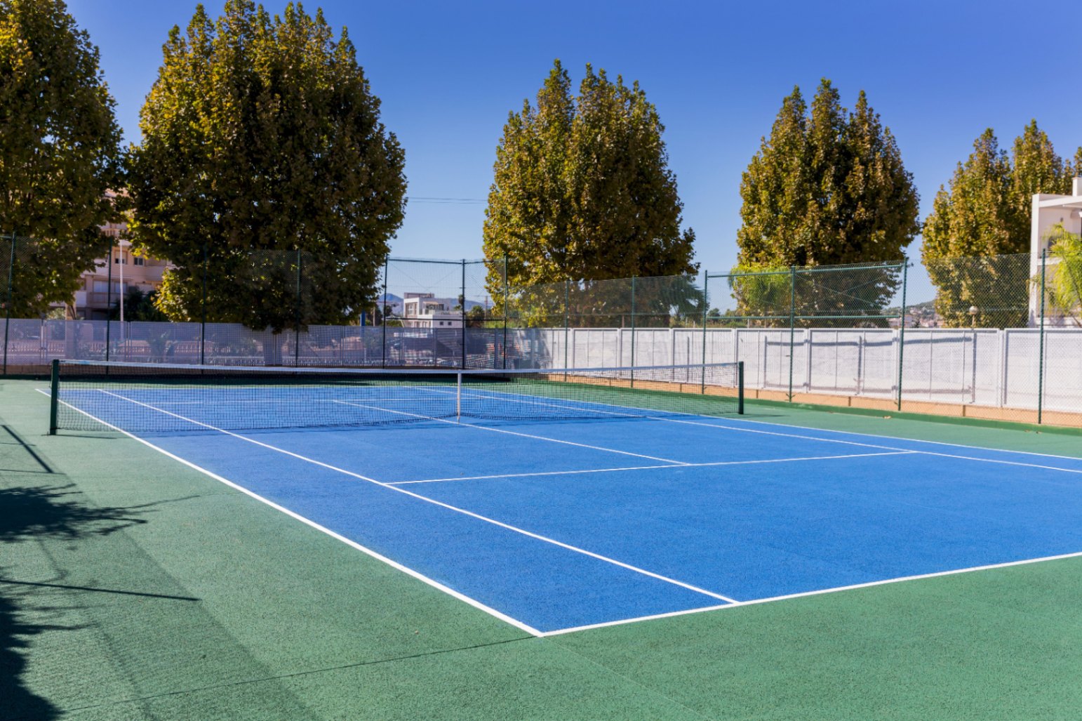 Pista de tenis exterior de superficie dura con zona de juego azul y perímetro verde, equipada con vallado perimetral y rodeada de árboles frondosos.
