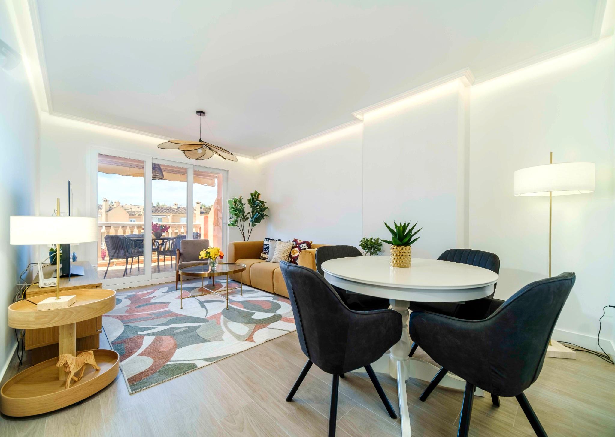 Salón comedor de planta abierta con sofá mostaza, mesa redonda blanca e iluminación LED perimetral. Puertas correderas dan acceso a una terraza equipada.