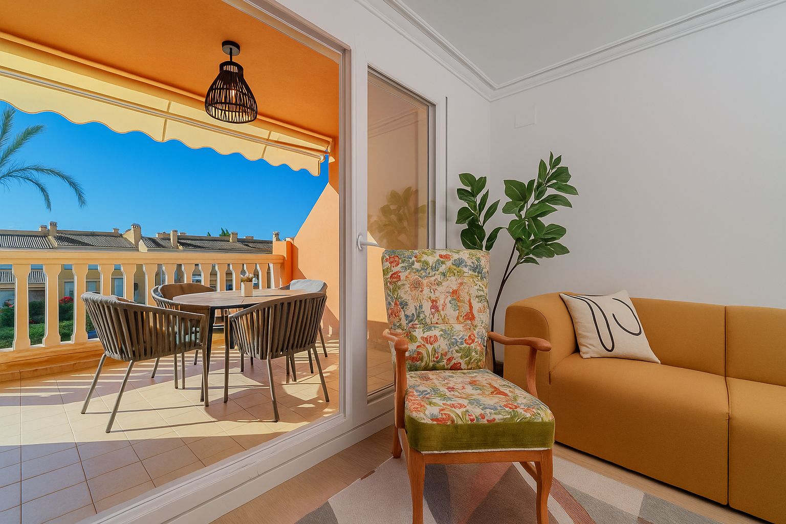Salón con sofá mostaza y sillón floral que conecta con una terraza privada equipada con comedor exterior, toldo amarillo y vistas despejadas.