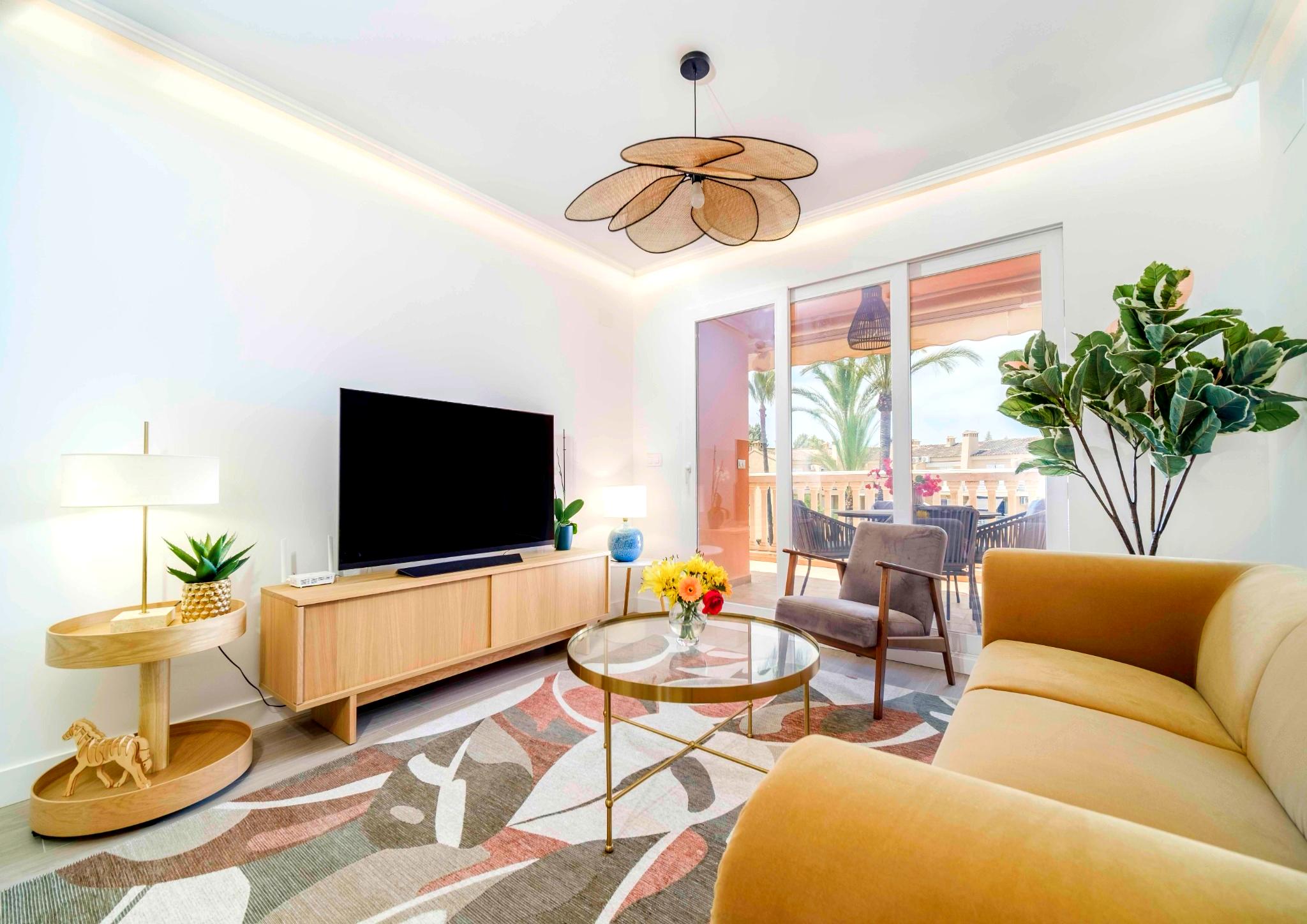Salón moderno con sofá mostaza, lámpara de techo tipo pétalo y puertas acristaladas que conectan con una terraza con vistas a palmeras.