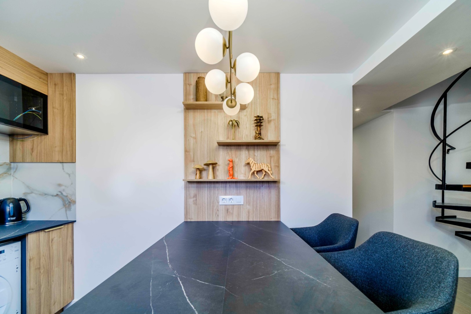 Zona de comedor moderna con mesa de efecto mármol oscuro, sillas tapizadas en azul y estantería de madera integrada bajo una lámpara de globos de diseño.