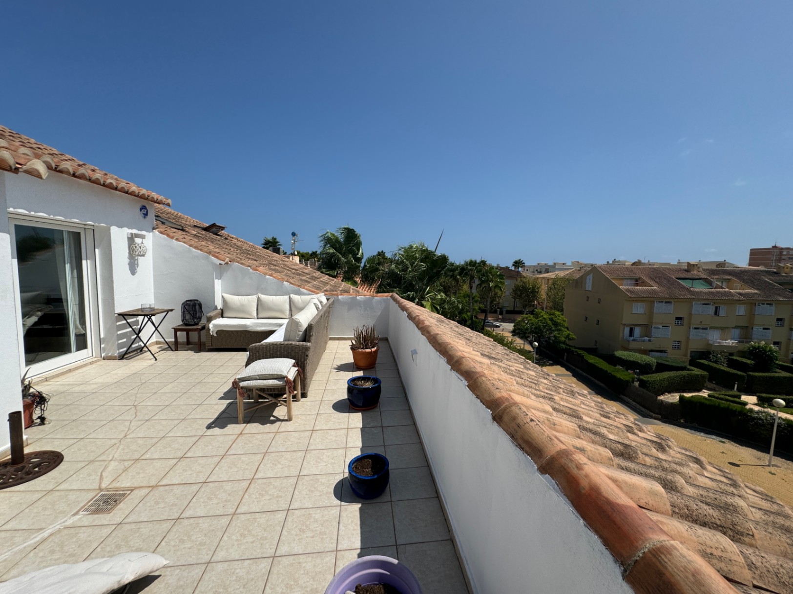 Amplia terraza en la azotea con suelo de baldosas claras, sofá de mimbre en L y vistas despejadas a una zona residencial con palmeras y tejados tradicionales.
