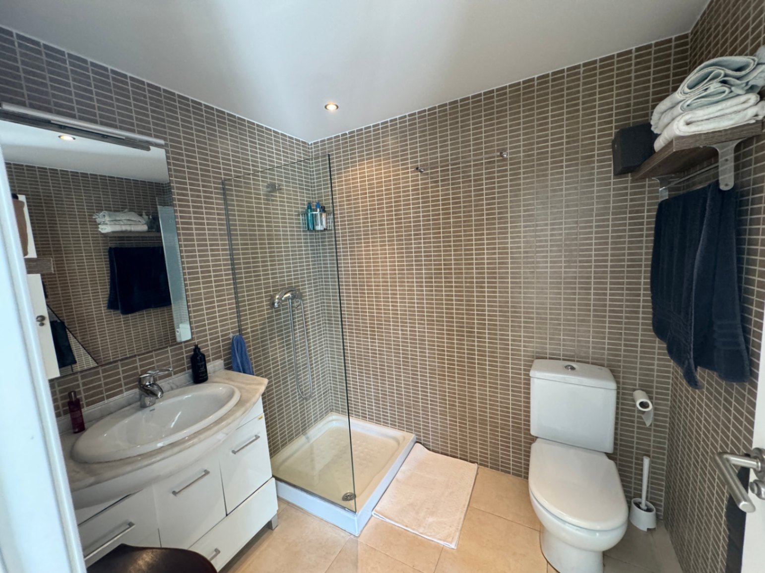 Baño con ducha de cristal, paredes de mosaico marrón y mueble de lavabo blanco con encimera de mármol e iluminación empotrada.