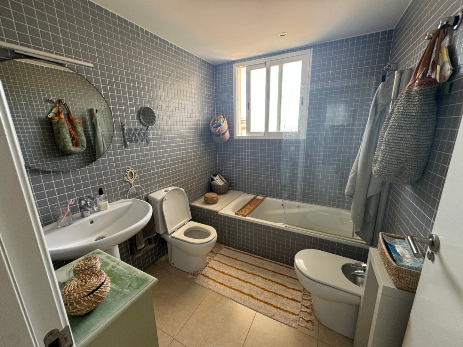 Baño con revestimiento de mosaico gris, lavabo de pedestal blanco, espejo circular y bañera con mampara de cristal bajo una ventana de luz natural.