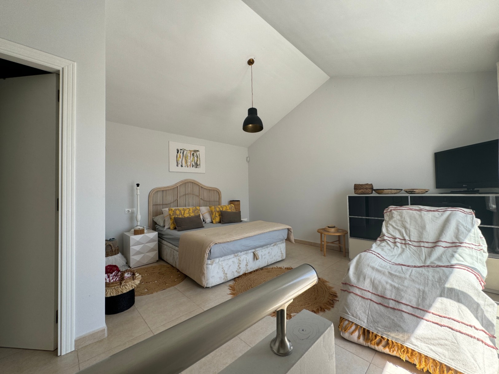 Dormitorio con techo abuhardillado, cabecero de mimbre y lámpara colgante negra. Incluye suelos cerámicos, alfombras de yute y una barandilla metálica en primer plano.