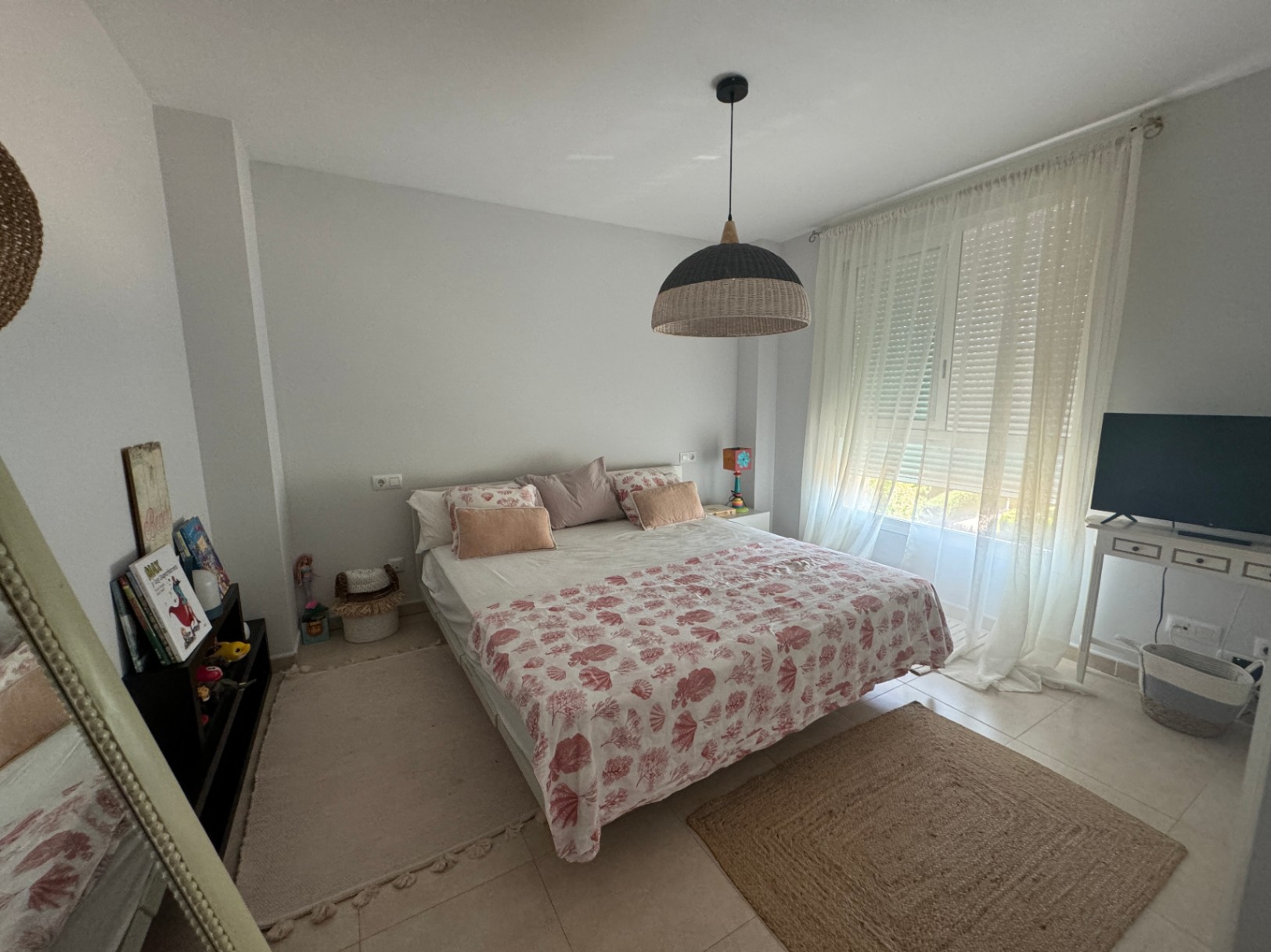 Dormitorio luminoso con cama doble, textiles de motivos marinos, lámpara de techo de fibras naturales y ventanal con cortinas traslúcidas.