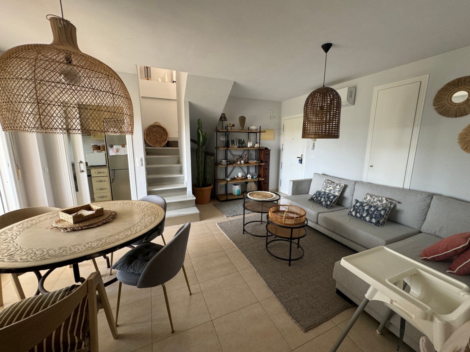 Salón-comedor de planta abierta con sofá modular gris, lámparas de mimbre colgantes y una mesa de comedor tallada junto a una escalera blanca minimalista.