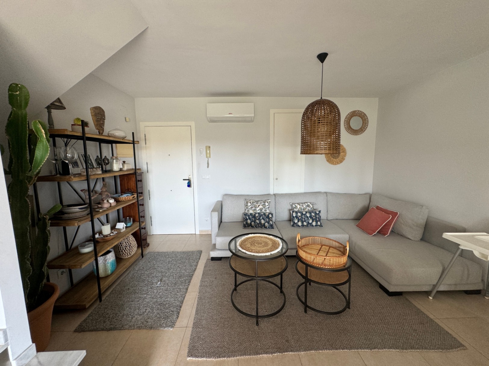 Salón con sofá en L gris, lámpara de techo de mimbre y estantería de madera y metal. El espacio cuenta con suelos cerámicos y una decoración de estilo mediterráneo.