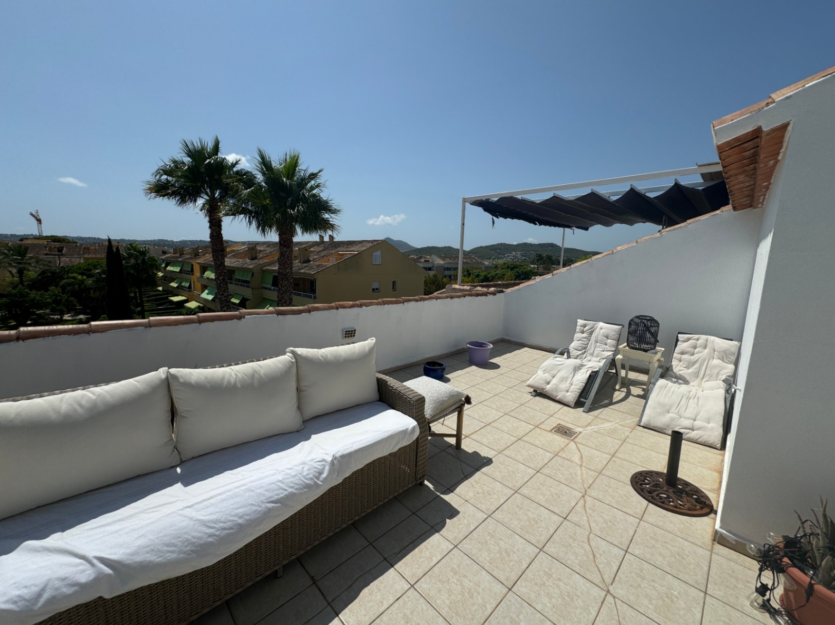 Terraza en la azotea con suelo cerámico, sofá de mimbre, tumbonas y toldo retráctil. Vistas despejadas a las palmeras y al entorno residencial.