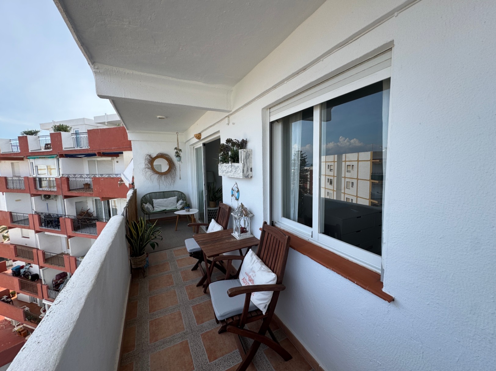 Balcón privado con suelo de baldosas cerámicas, mobiliario de madera plegable y una zona de descanso con sofá de mimbre y espejo decorativo.