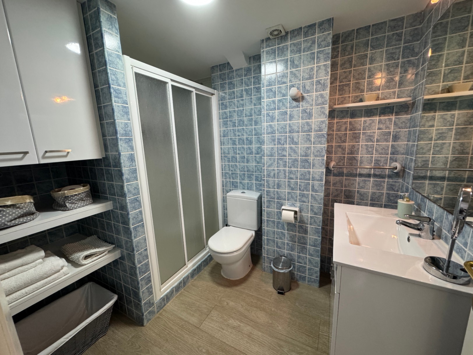 Baño con paredes de azulejos azules, lavabo integrado blanco y ducha con puertas correderas de cristal mate. Dispone de estanterías abiertas y suelo de efecto madera.