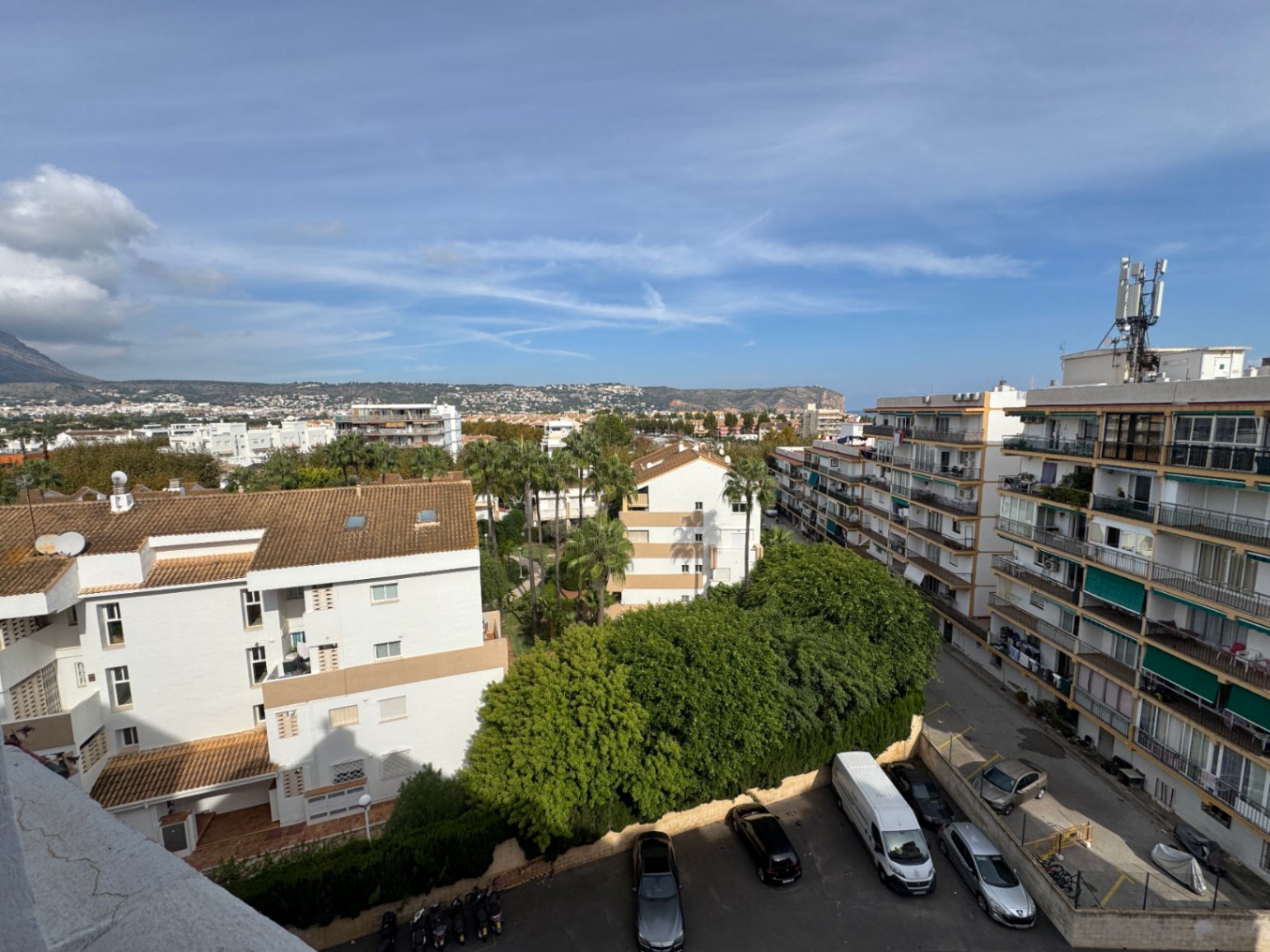 Vista elevada que destaca edificios residenciales, palmeras maduras y zona de aparcamiento, con colinas costeras visibles al fondo.