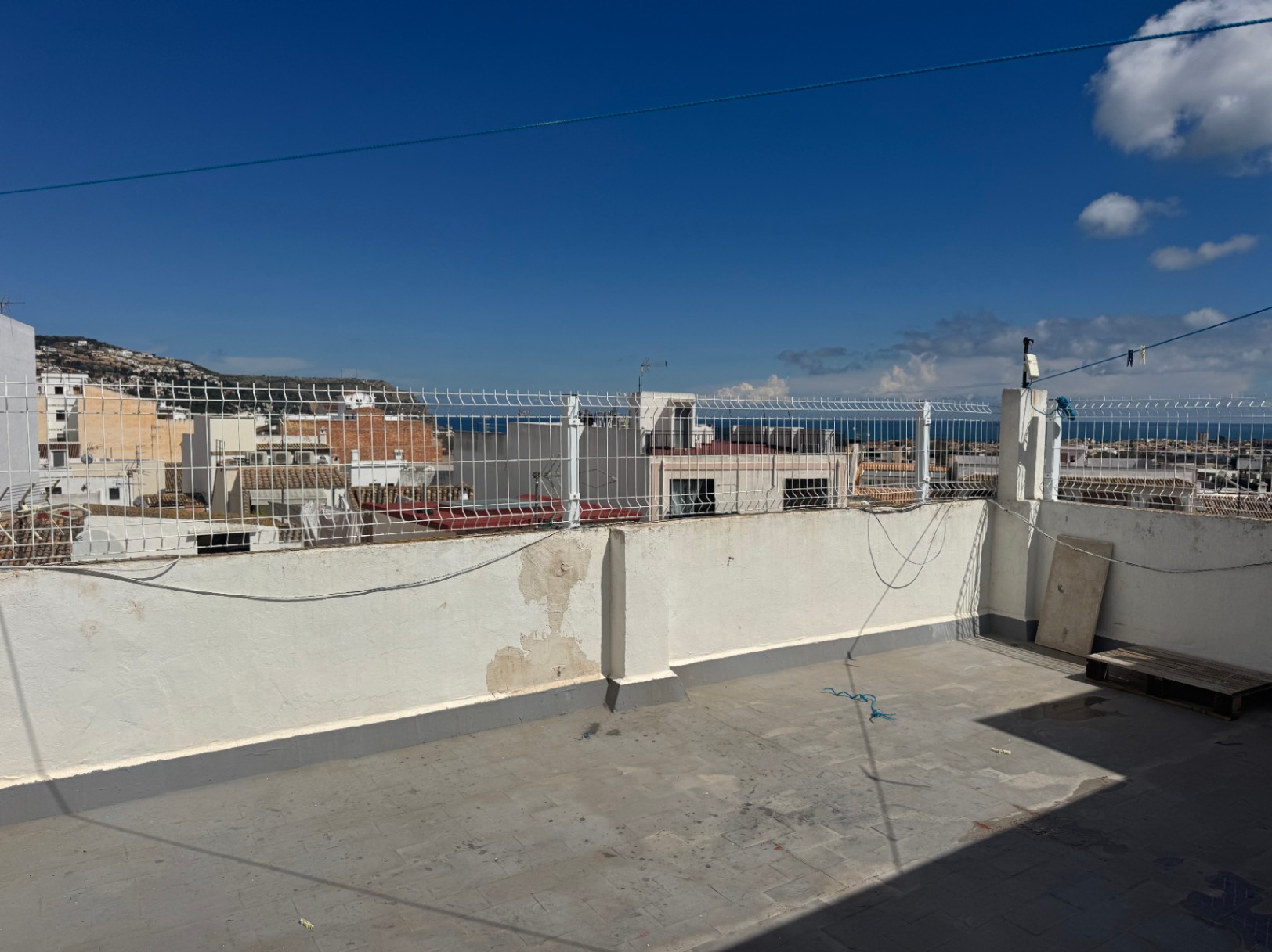 Terraza en la azotea con suelo de baldosas y muros blancos con valla metálica, ofreciendo vistas al entorno urbano, colinas y el mar en el horizonte.
