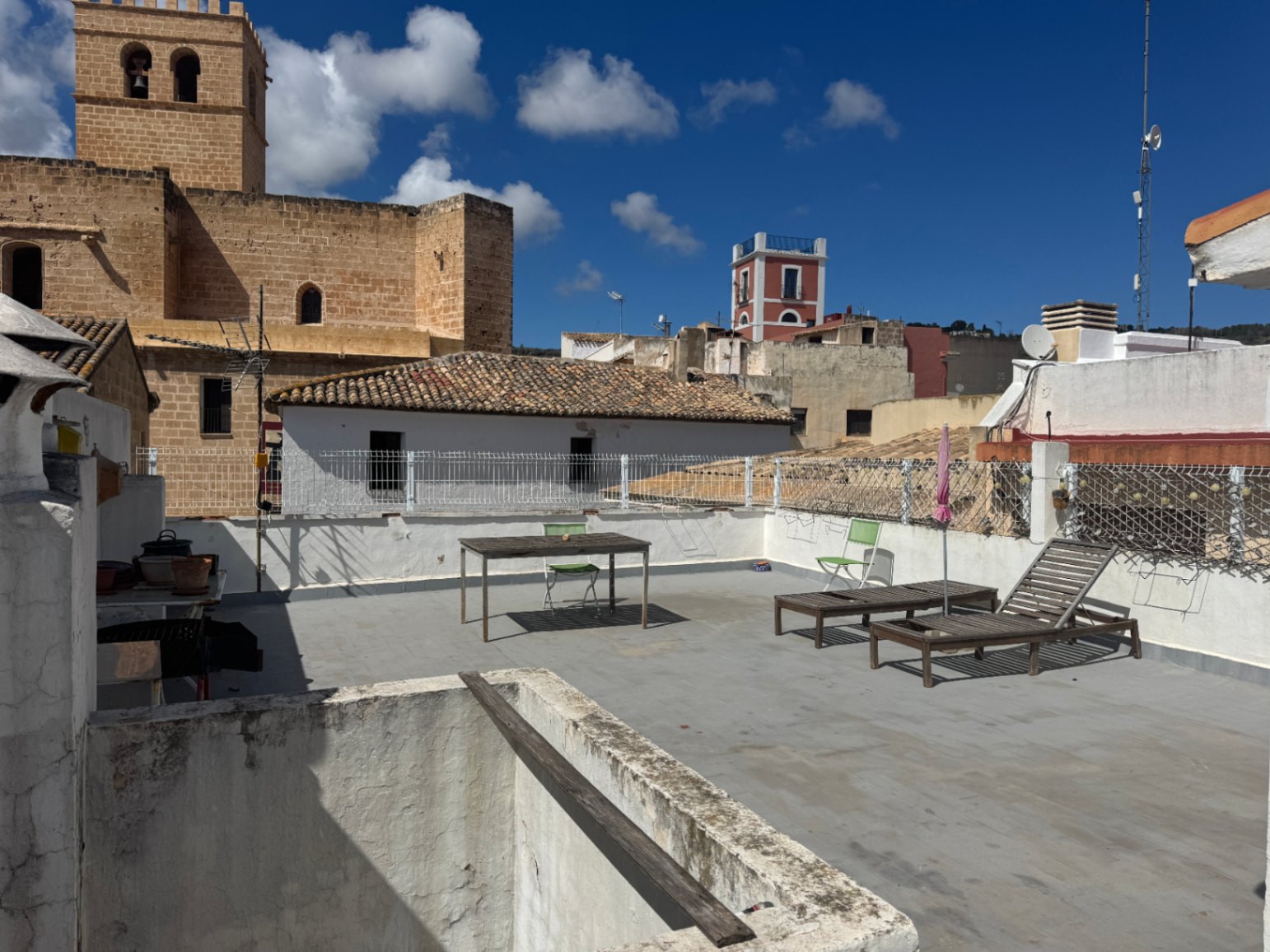 Terraza en la azotea con tumbonas de madera, mesa de comedor y vistas directas a una torre histórica de piedra y tejados tradicionales del pueblo.