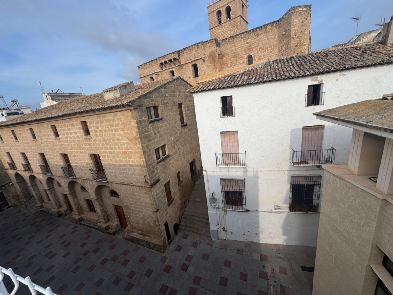 Vista de edificios históricos con arquerías de piedra y fachadas blancas frente a una plaza pavimentada, con una torre de iglesia al fondo.