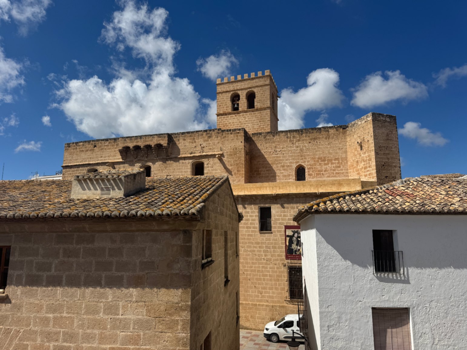 Vista de una iglesia fortificada de piedra tosca con campanario almenado, rodeada de casas tradicionales con tejados de teja cerámica bajo un cielo despejado.