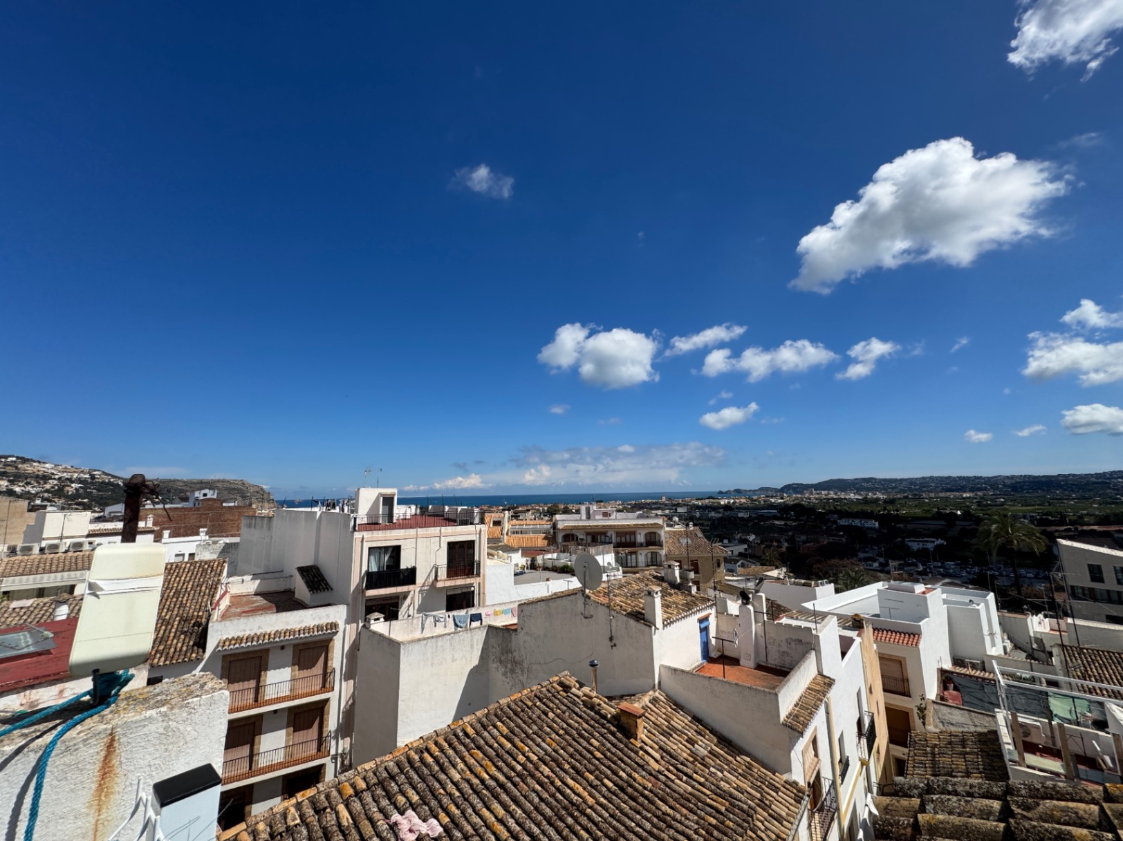 Vistas panorámicas elevadas sobre tejados de teja tradicional y fachadas blancas con el mar Mediterráneo en el horizonte bajo un cielo despejado.