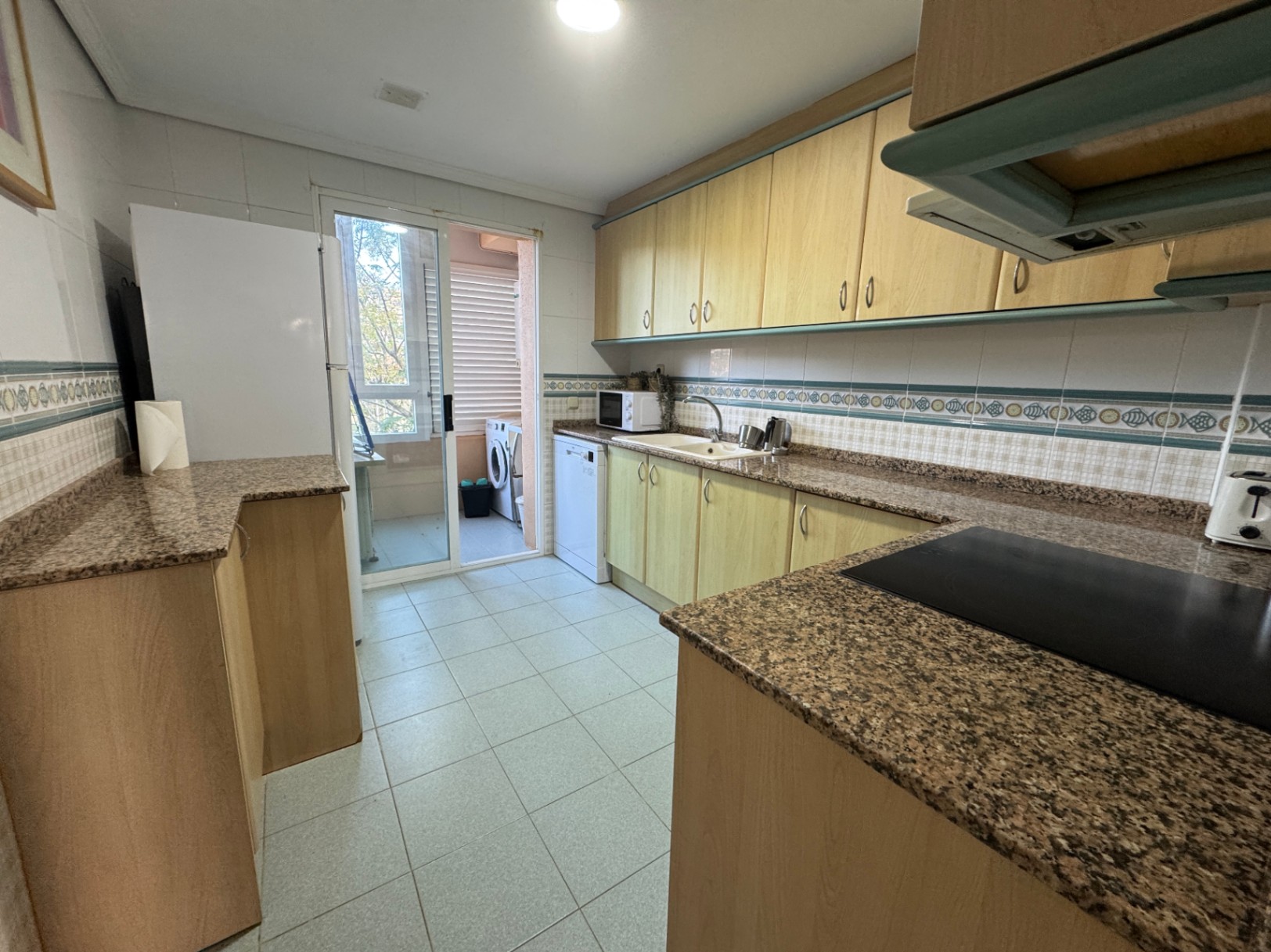 Cocina con armarios de madera clara, encimeras de granito y electrodomésticos blancos. Incluye acceso a un lavadero exterior mediante una puerta corredera.