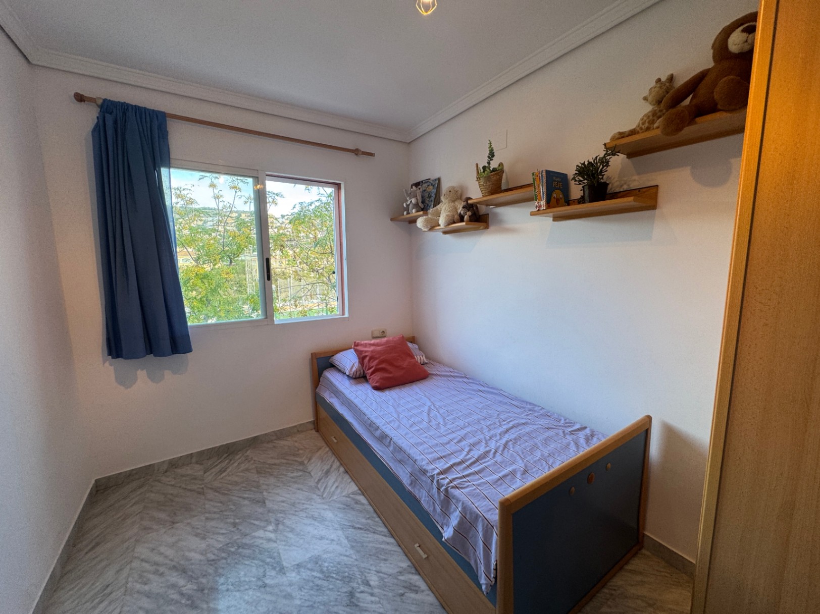 Dormitorio individual con suelos de mármol, estanterías de madera y ventana corredera con vistas. Incluye una cama con cajones de almacenamiento integrados.