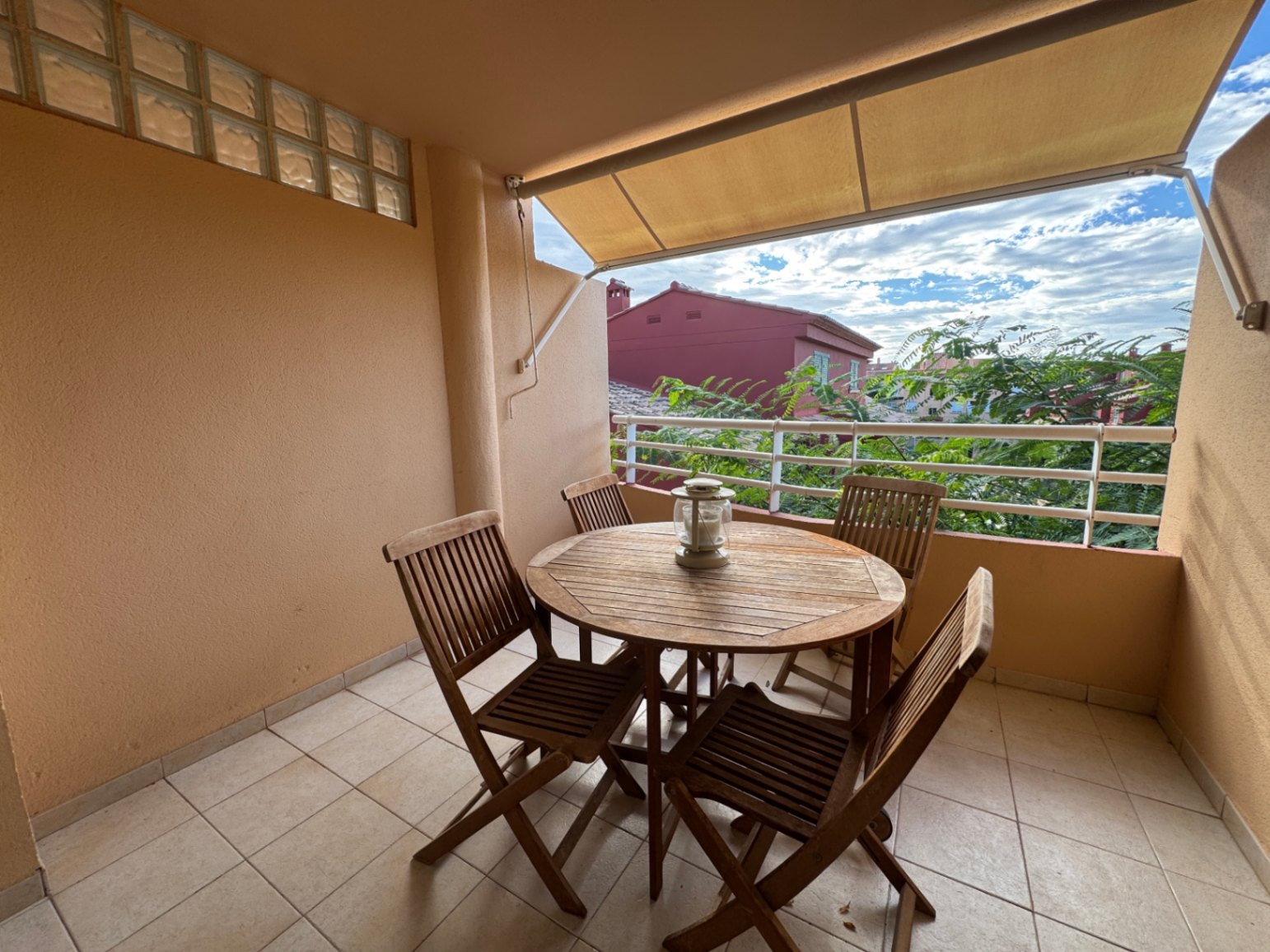 Terraza cubierta con conjunto de comedor de madera, suelo cerámico y toldo extensible con vistas a zonas verdes y edificios residenciales.